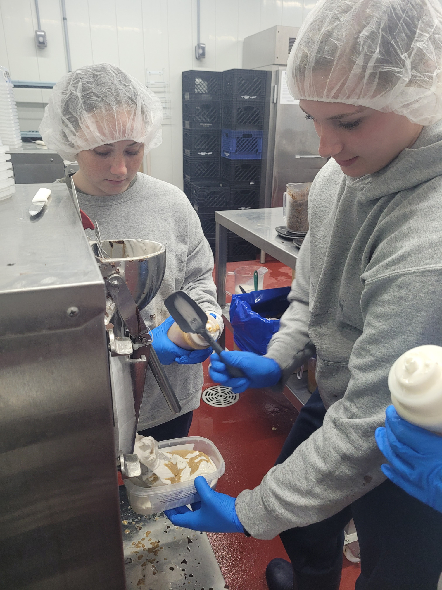 Two girls in grew sweatshirts and hair nets work on an ice cream batch.