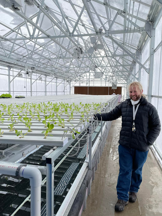 A man with blond hair stands in a greenhouse next to trays of hydroponic lettuce in its beginning stages.