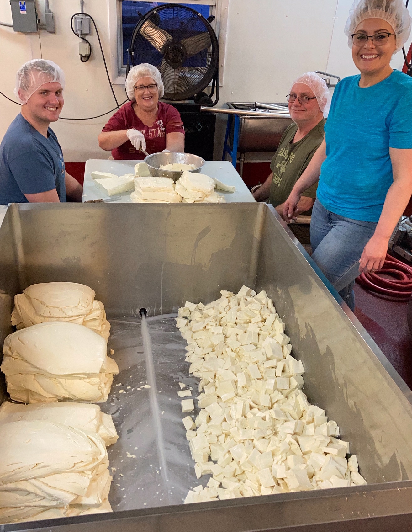 4 adults stand around a pile of cheese curds in the process of being cut up into small chunks.