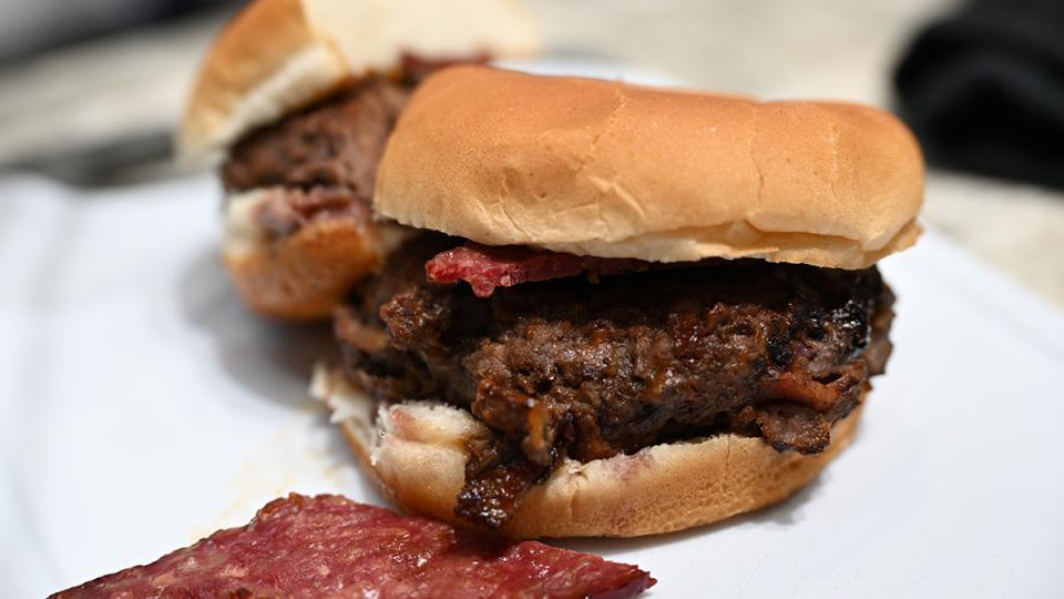 A bison burger and its bun are cut in half.  A piece of bison bacon sits in front of them both.