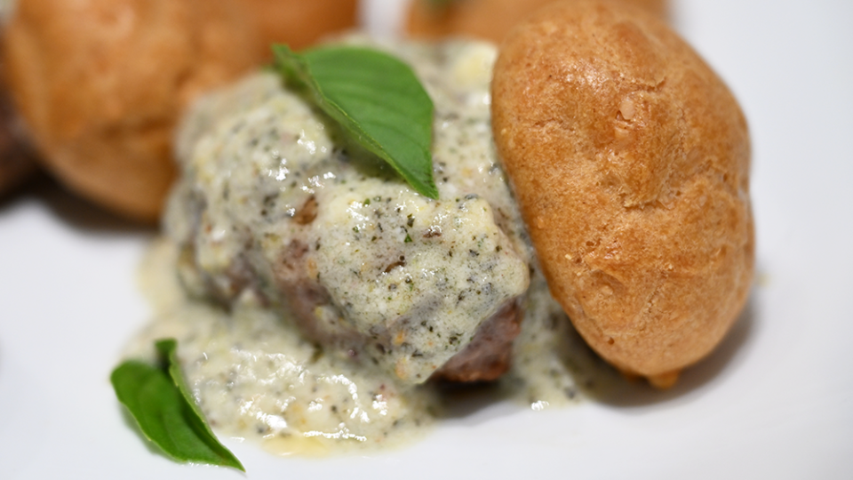 A lamb meatball is covered in sauce sitting next to a small gougère. Two miniature basil leaves complete the dish.