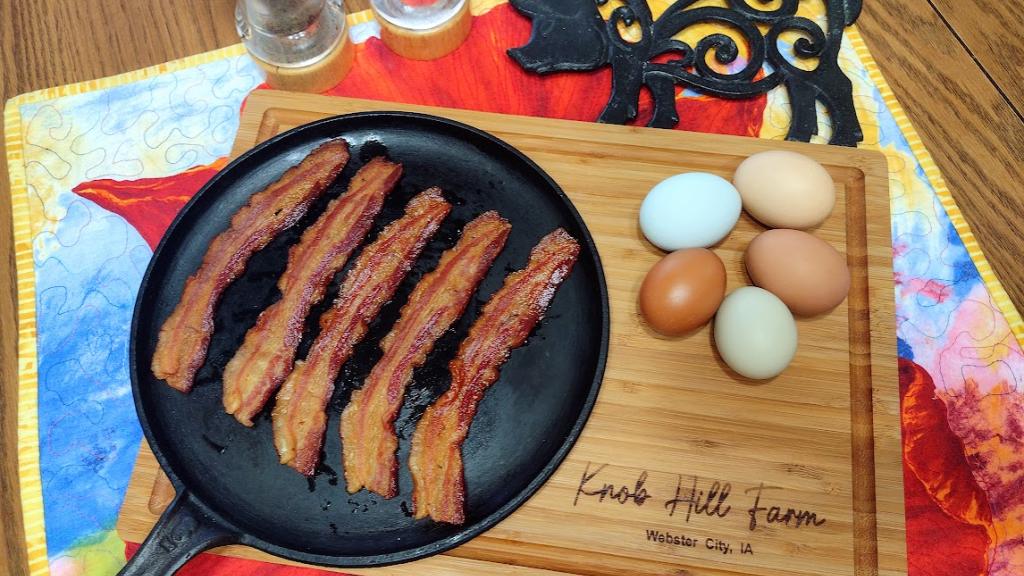 Cooked bacon on a cast iron skillet, set on a Knob Hill Farm wooden cutting board next to colorful eggs