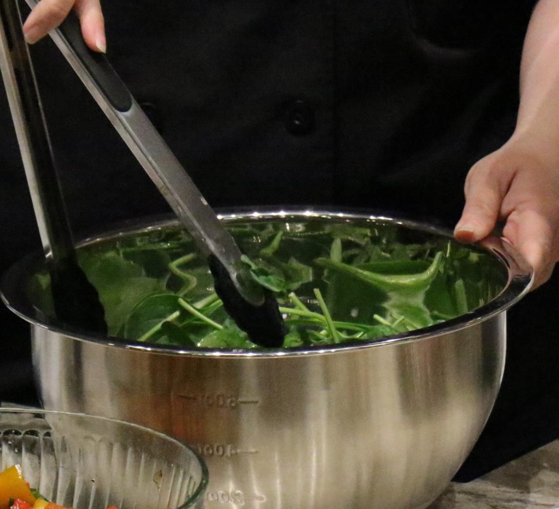 A pair of hands use tongs to mix up a bowl of spinach salad.