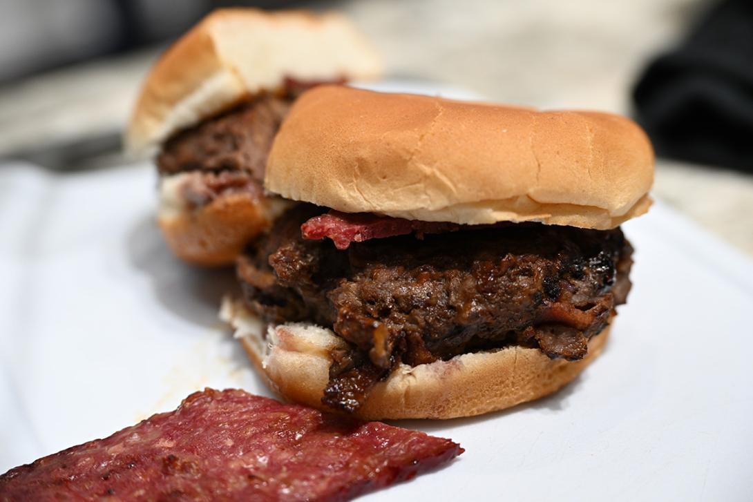 A bison burger and its bun are cut in half.  A piece of bison bacon sits in front of them both.